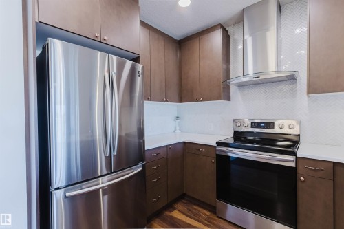 Kitchen featuring wood-finish cabinetry, stainless steel appliances, white countertops, a chevron-patterned backsplash, and recessed lighting - 205 1316 Windermere Way, Edmonton, AB - Indoor Photo Showing Kitchen With Stainless Steel Kitchen