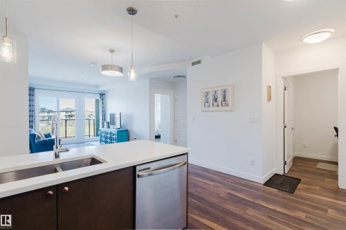 Modern kitchen island featuring a double stainless steel sink, stainless steel dishwasher, and white countertop - 205 1316 Windermere Way, Edmonton, AB - Indoor Photo Showing Kitchen