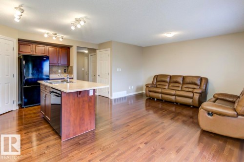 Open-concept living area featuring rich wood-finish flooring, a kitchen island with an integrated sink, dark wood cabinetry, and track lighting fixtures - 119 Rue Monique, Beaumont, AB - Indoor Photo Showing Kitchen With Double Sink