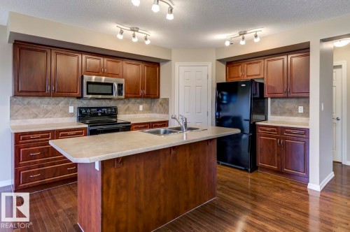 Kitchen featuring wood-finish flooring, a central island with a double sink, dark wood cabinetry, a tile backsplash, and track lighting - 119 Rue Monique, Beaumont, AB - Indoor Photo Showing Kitchen With Double Sink