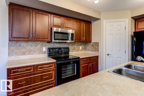 Kitchen featuring dark wood-finish cabinetry, light-toned countertops, a diagonal tile backsplash, stainless steel microwave, and a black electric range - 119 Rue Monique, Beaumont, AB - Indoor Photo Showing Kitchen With Double Sink