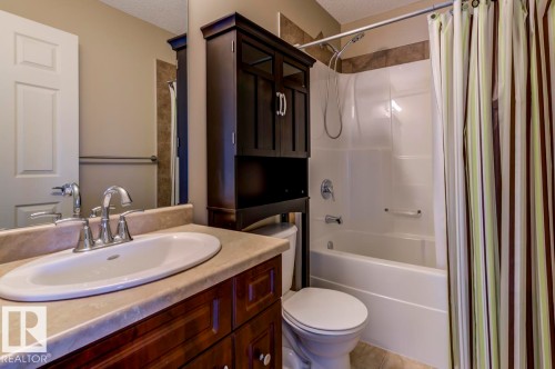 Bathroom featuring a built-in vanity with an oval sink and chrome faucet, a tub/shower combination with a shower head and separate spigot, a toilet, a dark wood-finish storage cabinet, and a large wall mirror - 119 Rue Monique, Beaumont, AB - Indoor Photo Showing Bathroom