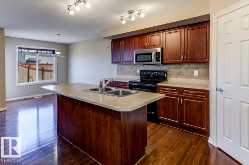 Kitchen featuring dark wood-finish cabinetry, stainless steel microwave, black electric range, and a light-toned countertop island with an integrated double sink - 119 Rue Monique, Beaumont, AB - Indoor Photo Showing Kitchen With Double Sink