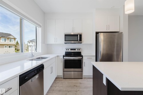 Modern kitchen featuring white cabinetry, stainless steel appliances, light-toned countertops, wood-finish flooring, and a large window - 3 11373 12 Avenue, Edmonton, AB - Indoor Photo Showing Kitchen With Stainless Steel Kitchen With Double Sink With Upgraded Kitchen