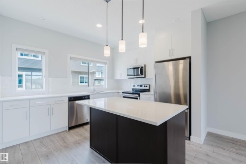 Modern kitchen featuring white cabinetry, a dark-toned island with a light countertop, stainless steel appliances, and light wood-finish flooring - 3 11373 12 Avenue, Edmonton, AB - Indoor Photo Showing Kitchen With Stainless Steel Kitchen With Upgraded Kitchen