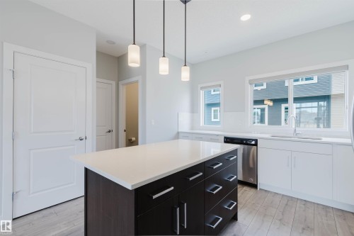 Modern kitchen featuring a dark wood-finish island with integrated storage, white perimeter cabinetry, stainless steel dishwasher, and a white countertop - 3 11373 12 Avenue, Edmonton, AB - Indoor Photo Showing Other Room