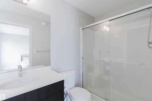 Bathroom featuring a dark wood-finish vanity with a white countertop and integrated sink, a frameless mirror, a toilet, and a glass-enclosed shower with sliding doors - 3 11373 12 Avenue, Edmonton, AB - Indoor Photo Showing Bathroom