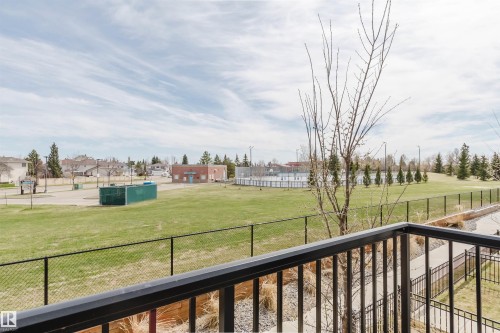 Balcony overlooking green space with black metal railing, offering expansive open field views, distant residential structures, and recreational facilities including a paved court and fenced rink - 3 11373 12 Avenue, Edmonton, AB - Outdoor
