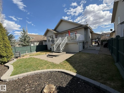 Rear exterior featuring a second-story deck with white railings and stairs, a paved patio, a grass lawn, and mature trees - 2335 Rutherford Way, Edmonton, AB - Outdoor