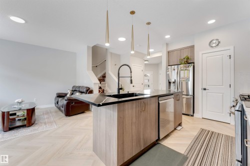 Open-concept kitchen featuring a waterfall island with a black sink and faucet, integrated dishwasher, light wood-finish cabinetry, and stainless steel appliances - 6404 27 Avenue, Edmonton, AB - Indoor Photo Showing Kitchen With Upgraded Kitchen