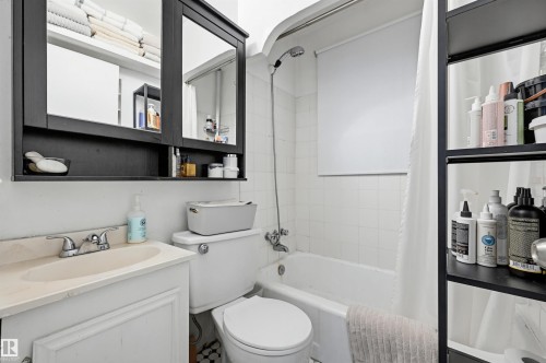 Bathroom featuring a white bathtub with subway tile surround, a white vanity with an integrated sink, a mirrored medicine cabinet, a white toilet, and a black shelving unit - 9816 93 Avenue, Edmonton, AB - Indoor Photo Showing Bathroom