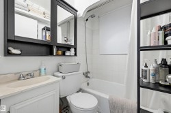 Bathroom featuring a white bathtub with subway tile surround, a white vanity with an integrated sink, a mirrored medicine cabinet, a white toilet, and a black shelving unit - 