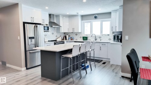 Contemporary kitchen featuring a large central island with a light-toned countertop, white cabinetry with sleek hardware, stainless steel appliances, a geometric pattern tile backsplash, and light wood-finish flooring - 3311 108 Street, Edmonton, AB - Indoor Photo Showing Kitchen With Upgraded Kitchen