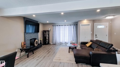 Living area featuring light wood-finish flooring, recessed ceiling lighting, a dark brick-faced fireplace, and a white paneled entry door with decorative glass - 3311 108 Street, Edmonton, AB - Indoor Photo Showing Living Room With Fireplace