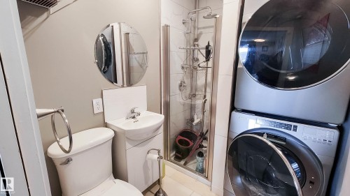 Compact bathroom featuring a glass-enclosed shower with a multi-function shower panel, a white pedestal sink with an integrated vanity, and a stacked washer and dryer unit - 3311 108 Street, Edmonton, AB - Indoor Photo Showing Laundry Room