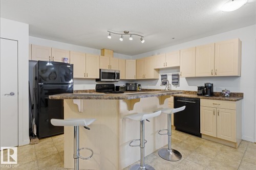 Kitchen featuring light-tone cabinetry, a peninsula with a raised breakfast bar, black appliances, and a track lighting fixture - 304 4312 139 Ave, Edmonton, AB - Indoor Photo Showing Kitchen