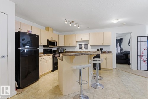 Kitchen featuring light wood-finish cabinetry, black appliances, track lighting, and an eat-at peninsula with stone-finish countertops - 304 4312 139 Ave, Edmonton, AB - Indoor Photo Showing Kitchen