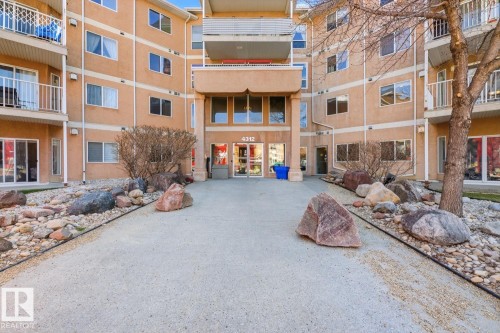 Multi-story stucco building featuring a central entry with a covered portico, multiple balconies with white railings, and an expansive concrete pathway - 304 4312 139 Ave, Edmonton, AB - Outdoor With Balcony