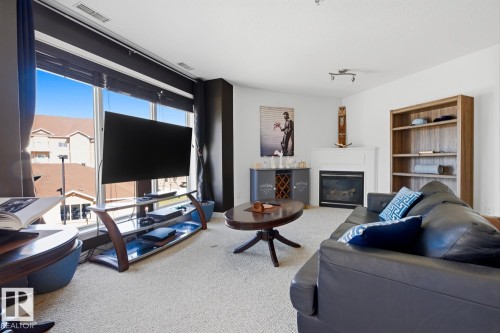 Living room featuring a large picture window with blinds, a corner fireplace with a white mantel, and light-colored carpeting - 304 4312 139 Ave, Edmonton, AB - Indoor With Fireplace
