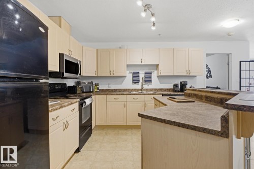 Kitchen featuring light wood-finish cabinetry, dark countertops, black range, and stainless steel microwave - 304 4312 139 Ave, Edmonton, AB - Indoor Photo Showing Kitchen With Double Sink