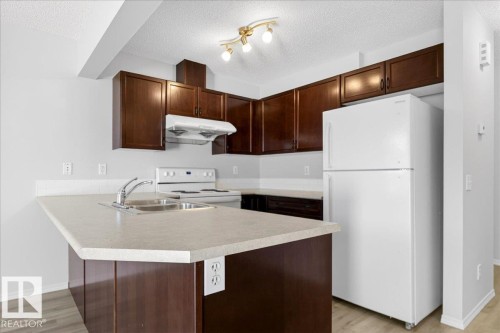 Kitchen featuring dark wood-finish cabinetry, light-toned countertops, a white appliance package, a stainless steel sink with a chrome faucet, and wood-finish flooring - #7 18004 89 St Nw, Edmonton, AB - Indoor Photo Showing Kitchen With Double Sink