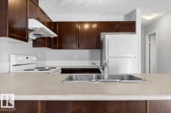 Kitchen featuring rich wood-finish cabinetry, light-toned countertops, a stainless steel double-basin sink with a chrome faucet, a white electric range, and a white refrigerator - 