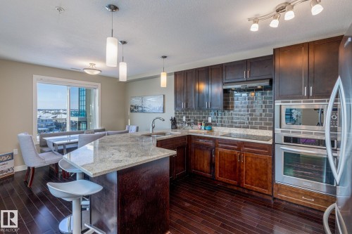 Modern kitchen featuring a granite-topped island with breakfast bar seating, dark wood-finish cabinetry, stainless steel appliances, and a subway tile backsplash - 513 1230 Windermere Way, Edmonton, AB 