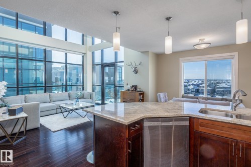Open-concept living area featuring a kitchen island with stone countertop and integrated stainless steel dishwasher, rich wood-finish cabinetry, dark wood-finish flooring, and a two-story window wall - 513 1230 Windermere Way, Edmonton, AB 