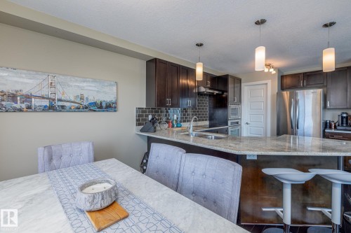 Kitchen featuring dark wood cabinetry, a granite-finish island with seating, stainless steel appliances, a tile backsplash, and recessed and pendant lighting - 513 1230 Windermere Way, Edmonton, AB 