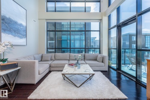 Living area featuring soaring two-story windows with dark framing, rich wood-finish flooring, and a private balcony with glass railing - 513 1230 Windermere Way, Edmonton, AB 