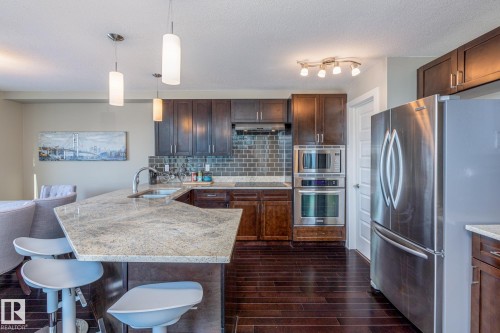 Kitchen featuring dark wood cabinetry, stainless steel appliances, a tile backsplash, and a light-toned countertop with an integrated sink - 513 1230 Windermere Way, Edmonton, AB 