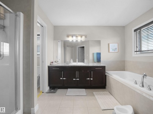 Bathroom featuring a spacious built-in tub, a dark wood-finish double vanity with a light-toned countertop, a large mirror, and tiled flooring - 3247 15 Avenue Nw, Edmonton, AB 