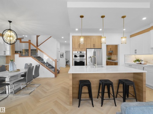 Open-concept kitchen and dining area featuring herringbone wood-finish flooring, a central island with a quartz-finish countertop, stainless steel appliances, and a staircase with wood railings and glass paneling - 209 Crystal Creek Dr Nw, Leduc, AB - Indoor