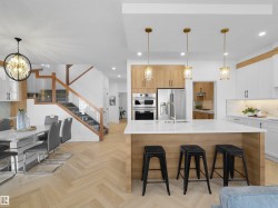 Open-concept kitchen and dining area featuring herringbone wood-finish flooring, a central island with a quartz-finish countertop, stainless steel appliances, and a staircase with wood railings and glass paneling - 