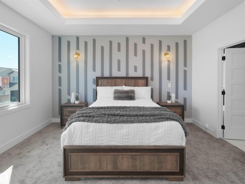 Recessed tray ceiling with ambient lighting, accent wall with geometric pattern, two wall sconces with gold-tone fixtures, light gray carpeting, and a white framed window - 209 Crystal Creek Dr Nw, Leduc, AB - Indoor Photo Showing Bedroom