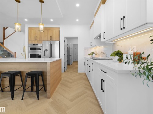 Contemporary kitchen featuring a herringbone wood-finish floor, white shaker cabinetry with matte black hardware, integrated stainless steel appliances, a white countertop, and a kitchen island with a golden faucet - 209 Crystal Creek Dr Nw, Leduc, AB - Indoor Photo Showing Kitchen With Upgraded Kitchen
