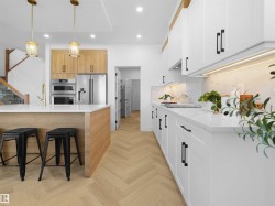 Contemporary kitchen featuring a herringbone wood-finish floor, white shaker cabinetry with matte black hardware, integrated stainless steel appliances, a white countertop, and a kitchen island with a golden faucet - 