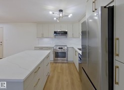 Open-concept kitchen featuring an oversized island with a quartz countertop, wood-finish flooring, stainless steel appliances, light gray cabinetry, and a contemporary ceiling fixture - 