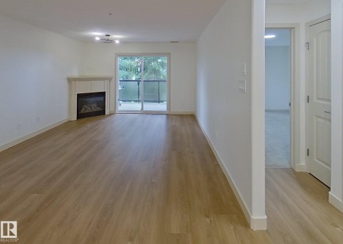 Spacious living area featuring wood-finish flooring, a white mantel fireplace, and a sliding glass door opening to a private balcony with glass railing - 353 2096 Blackmud Creek Drive, Edmonton, AB - Indoor Photo Showing Living Room With Fireplace