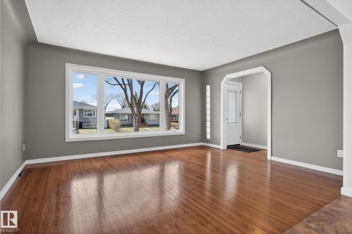 Spacious living area featuring wood-finish flooring, a large picture window, neutral gray wall paint, and white trim throughout - 10409 83 Street, Edmonton, AB - Indoor Photo Showing Other Room