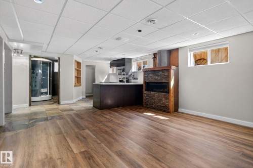 Spacious open concept room featuring wood-finish flooring, a stone-faced fireplace with a wood mantle, a kitchen area with dark cabinetry, a built-in shower enclosure, and recessed lighting - 10409 83 Street, Edmonton, AB - Indoor