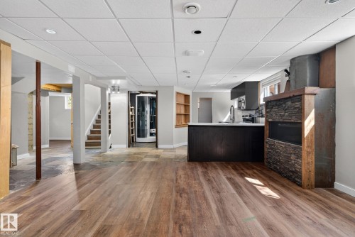 Spacious room with wood-finish flooring, a built-in fireplace featuring stone and wood accents, and a kitchen area with dark cabinetry and light countertops - 10409 83 Street, Edmonton, AB - Indoor