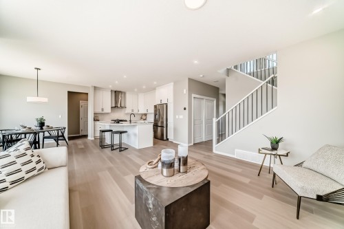 Open-concept living space featuring wood-finish flooring, a contemporary kitchen with white cabinetry and stainless steel appliances, a dining area with a modern chandelier, and a staircase with metal balusters - 1603 206 Street, Edmonton, AB - Indoor Photo Showing Living Room
