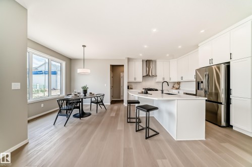 Open-concept kitchen and dining area featuring light wood-finish flooring, white cabinetry with black hardware, stainless steel appliances, a kitchen island with seating, and a dining area with ample natural light from large windows - 1603 206 Street, Edmonton, AB - Indoor