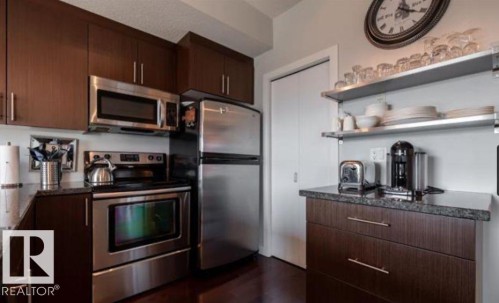 Kitchen featuring dark wood-finish cabinetry, stainless steel appliances, dark stone-finish countertops, and open shelving - 702 10055 118 Street, Edmonton, AB - Indoor Photo Showing Kitchen