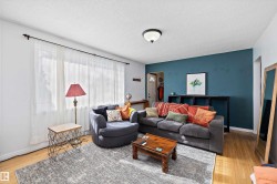 Living area featuring wood-finish flooring, a contemporary ceiling light fixture, a prominent accent wall, and a large window with sheer white curtains - 