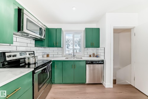 Modern kitchen featuring green cabinetry with gold-tone hardware, stainless steel appliances, white subway tile backsplash, and light wood-finish flooring - 8712 89 Avenue, Edmonton, AB 