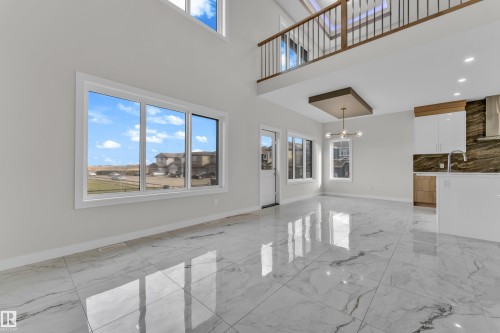 Two-story living area with high ceilings and large windows, featuring white marble-look flooring, a kitchen island with an integrated sink, and an upper-level balcony with a wood and metal railing - 12603 44 Street, Edmonton, AB - Indoor Photo Showing Other Room