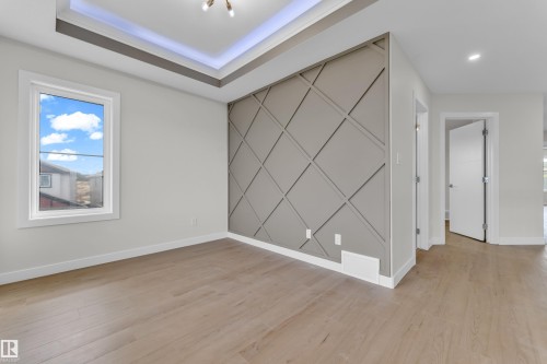 Spacious room featuring wood-finish flooring, a decorative accent wall with geometric paneling, a tray ceiling with recessed lighting, and a window with white trim - 12603 44 Street, Edmonton, AB - Indoor Photo Showing Other Room