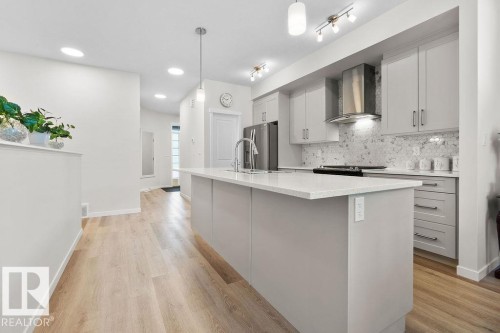 Modern kitchen featuring a central island with a light-toned countertop, light gray cabinetry, stainless steel appliances, and a hexagonal tile backsplash - 3623 6 Street, Edmonton, AB - Indoor Photo Showing Kitchen With Upgraded Kitchen